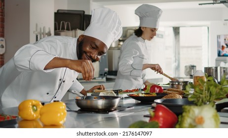 African american cook pouring shredded parmesan cheese on top of restaurant dish in frying pan. Male chef in uniform cooking professional gourmet meal with grated cheddar and ingredients in kitchen. - Powered by Shutterstock