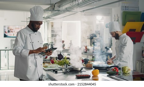 African American Cook Searching Gourmet Recipe On Digital Tablet, Using Device With Technology To Prepare Professional Food Dish. Male Chef In Uniform Cooking Gastronomic Meal From Website.