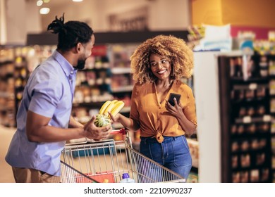 African American Couple Choosing Products Using Phone During Grocery Shopping In Modern Supermarket. Selective Focus