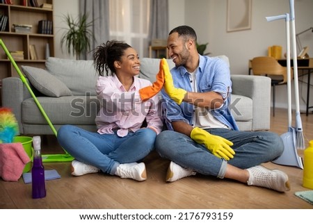 African American Couple Giving High Five Cleaning House Doing Housework Together Sitting On Floor In Modern Living Room At Home. Family Lifestyle, House Chores Concept