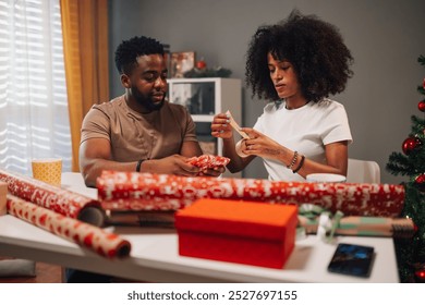 An african american couple happily wraps Christmas gifts with red paper while seated at a table. The atmosphere is joyful and warm, enhanced by a beautifully decorated Christmas tree. - Powered by Shutterstock