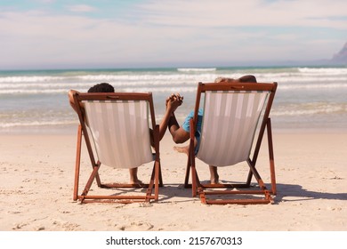 African american couple holding hands while relaxing on deckchairs at beach against cloudy sky. 