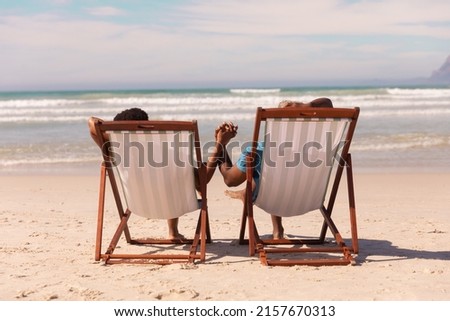 African american couple holding hands while relaxing on deckchairs at beach against cloudy sky. 