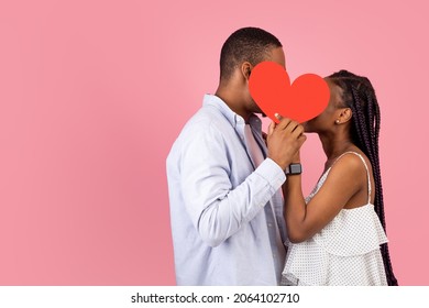 African American Couple In Love Kissing, Holding And Hiding Behind Red Paper Heart Card, Covering Their Faces For Privacy, Isolated On Pink Background. St. Valentines Day Concept, Free Copy Space