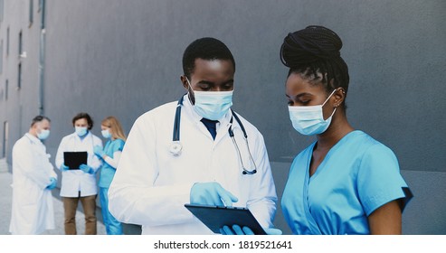 African American Couple Of Man And Woman, Doctors Colleagues In Medical Masks Working And Using Tablet Device. Male And Female Physicians Talking, Tapping And Scrolling On Gadget Computer.