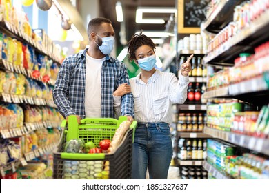 African American Couple In Protective Masks In Supermarket Doing Grocery Shopping, Buying Food Essentials Walking With Shop Cart In Groceries Store Indoors. Family Choosing Food Products On Shelves