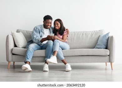 African American couple sharing social media on cellphone, happy partners sitting on the sofa, male showing gadget. Smiling young black guy embracing lady looking at smartphone screen, free copy space - Powered by Shutterstock