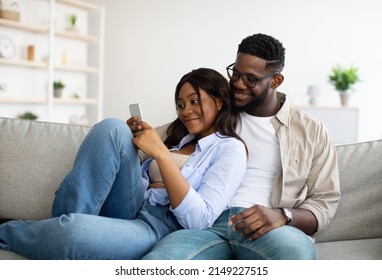 African American Couple Sharing Social Media On Cellphone, Happy Family Sitting On The Sofa, Using Gadget. Smiling Young Black Man In Glasses And Braces Embracing Woman Looking At Smartphone Screen