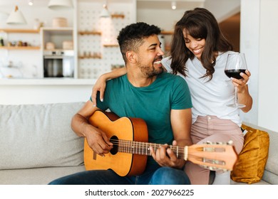 The African American couple is spending romantic moments together. The man is playing the guitar with his beloved girlfriend. A woman is sitting next to him on the couch and drinking red wine.
