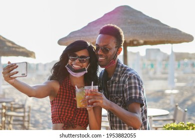 African american couple taking selfie in beach party - Young friends with face mask drinking cocktail in holiday - Coronavirus summer - Powered by Shutterstock