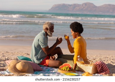 African american couple toasting juice while sitting with food on blanket against sea at sunset. happy, nature, unaltered, love, togetherness, retirement, picnic, enjoyment and holiday concept. - Powered by Shutterstock
