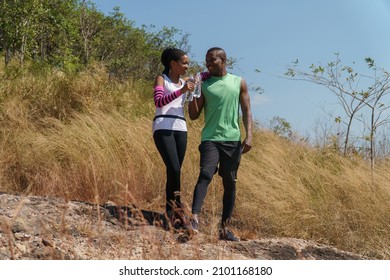 African American Couple Trail Running Take A Break Drinking Water In Meadow On Mountain Outdoors. Sport Adventure . Black Fit Woman And Man Runner Jogging On Country Path Or Cross-country In Summer