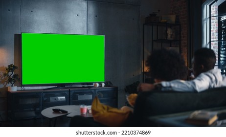 African American Couple Watching TV With Green Screen Mockup Display. They Are Sitting On A Couch In Their Cozy Living Room And Eating Popcorn, Enjoying Streaming Service Movie Night. Back View.