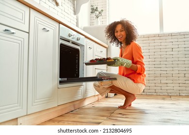 African American cute woman with curly fluffy hair taking out a baking sheet with baked muffins from the oven. - Powered by Shutterstock