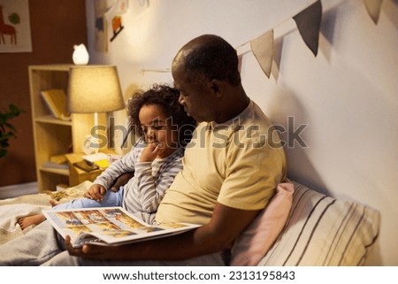 African American dad reading fairy tale to his child, they sitting on bed and watching pictures in book