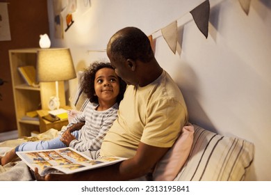 African American dad reading tale to his child before sleep, they watching pictures and talking to each other