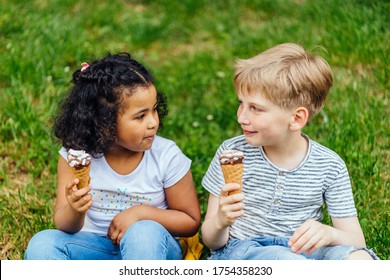 African American Dark Skinned Little Girl With Older Preteenager Blond Boy With Ice Cream, Scheming And Conspiring, Thinking Devious Tricks And Cheats, Cunning And Betraying Against Green Lawn Outdoor