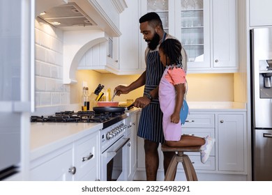 African american daughter looking at father cooking pancakes on stove in kitchen. Unaltered, lifestyle, family, love, togetherness, childhood, food, preparation and learning concept.