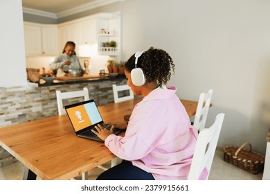 African american daughter using laptop while mother cooking in background at home - Family lifestyle concept - Main focus on headphones - Powered by Shutterstock