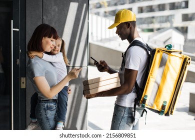 African American delivery man with boxes of hot pizza. Young smiling woman with a girl at the door, pay by card for online order. Fast Home Delivery. Beautiful family mother, daughter.