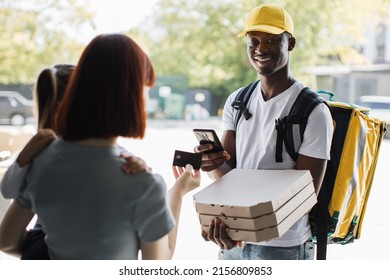 African American delivery man with boxes of hot pizza. Young smiling woman with a girl at the door, pay by card for online order. Fast Home Delivery. Beautiful family mother, daughter.
