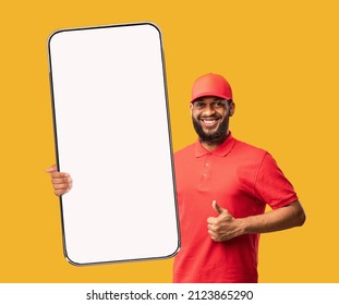African American Delivery Man Demonstrating Huge Cellphone With Blank Screen And Gesturing Thumb Up, Approving Shipping Application Over Yellow Background, Mockup