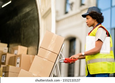 African American Delivery Woman With Hand Truck Unloading Packages From A Truck.