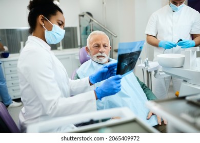 African American Dentist Showing To Senior Man His Orthopantomogram During Dental Appointment.