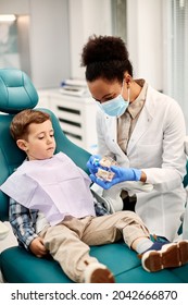 African American Dentist Teaching Little Boy How To Brush His Teeth During Dental Check-up At Dentist's Office.