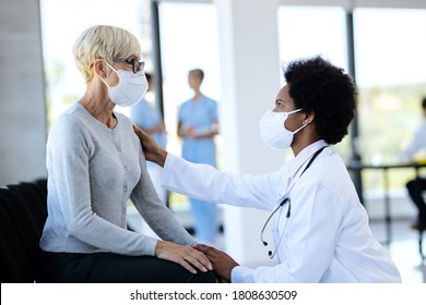 African American Doctor Consoling Her Mature Patient In Waiting Room At Medical Clinic. Both Of Them Are Wearing Protective Face Masks. 