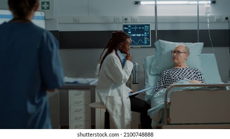 African American Doctor Consulting Sick Elder Patient In Hospital Ward While Nurse Writes Prescription On Computer. Old Man With Oximeter And Nasal Oxygen Tube Receiving Treatment