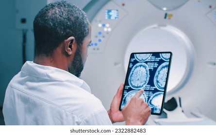 African American doctor examine scan attentively. Medical worker shooted from back. Male doctor examines MRI scan. Multicultural doctor is scrolling screen of tablet with scans of brain. - Powered by Shutterstock