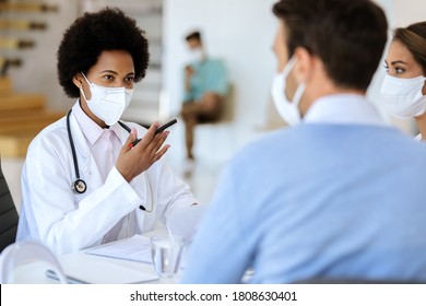 African American doctor having an appointment with a couple and wearing protective face mask while communicating with them at doctor's office. 
