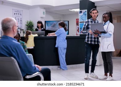 African American Doctor Holding Laptop With Mri Scan Results Talking With Asian Patient In Busy Private Clinic Waiting Room. Medic Having Conversation With Young Man Checking Appointment.