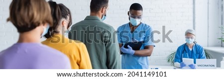 african american doctor in medical mask writing on clipboard near patients and blurred asian colleague, banner