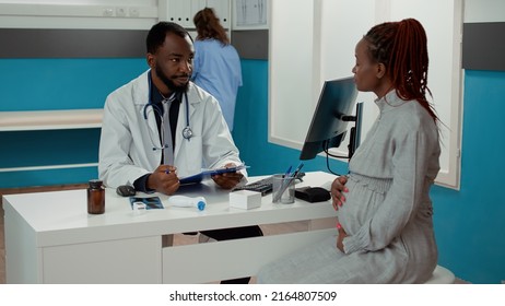 African american doctor and patient expecting child meeting in cabinet at checkup visit. Male physician and pregnant woman having conversation about childbirth at consultation appointment. - Powered by Shutterstock