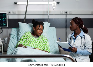 African american doctor and patient talking in hospital ward about diagnosis and treatment for teenager in bed. Black specialist woman with stethoscope consulting sick young adult - Powered by Shutterstock