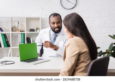 African American Doctor Pointing With Pen At Laptop With Green Screen Near Brunette Woman