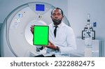 African American doctor is posing for camera. Mature man is showing screen of chroma key tablet. Multicultural doctor is smiling and looking at camera. Medical worker at background of MRI capsule.