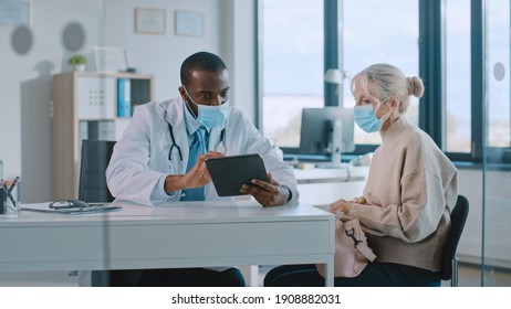 African American Doctor In Protective Mask Is Reading Medical History Of Senior Female Patient During Consultation In A Health Clinic. Physician Using Tablet Computer In Hospital Office.