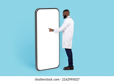 African American Doctor Standing Near Big Blank Smartphone And Touching White Screen, Young Black Male Physician Browsing New Mobile Application While Standing On Blue Background, Collage, Mockup