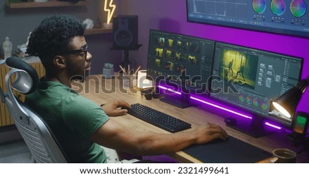 African American editor makes color correction, works at home studio. Action movie footage and program interface on multi-monitor computer and big digital screen. Concept of film post production.