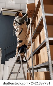 African american employee standing on ladder in storehouse, preparing customers orders using cardboard boxes. Small business worker wearing overall doing merchandise quality control in warehouse