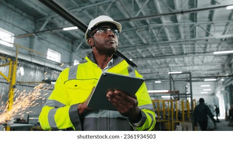 African American engineer in hard hat and protective vest is looking through workshop. Man is holding gadget and gesturing at construction site. Employee on background of welding sparks flying. - Powered by Shutterstock