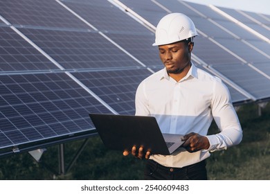 African American engineer wearing a hard hat uses a laptop in front of solar panels. This image captures the essence of renewable energy and technology integration in an outdoor setting. - Powered by Shutterstock