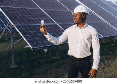 African American Engineer in a white hard hat holding a light bulb, standing near solar panels outdoors. Represents clean energy, innovation, and sustainable technology in a sunny landscape. - Powered by Shutterstock