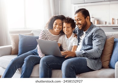 African American family consisting of a father, mother, and two children sitting comfortably on a couch. They are all focused on a laptop screen, engaged in an activity together. - Powered by Shutterstock