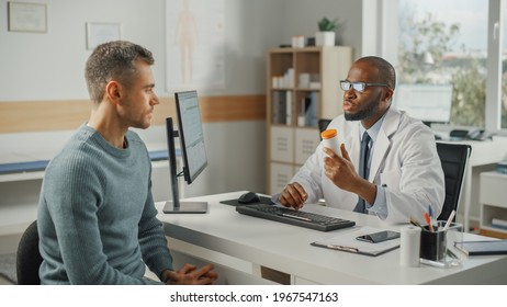 African American Family Doctor Is Talking With Young Male Patient During Consultation In A Health Clinic. Black Physician In Lab Coat And Glasses Sitting Behind A Computer Desk In Hospital Office.