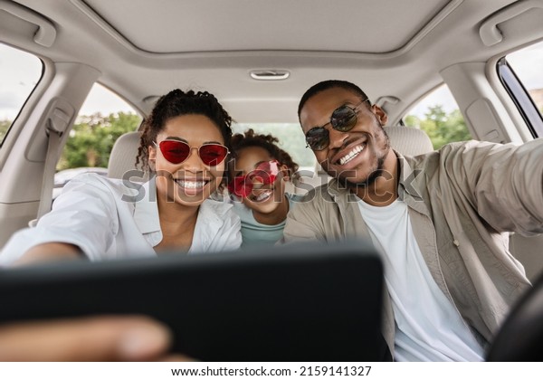 African American Family Making Selfie In New Car On\
Smartphone. Happy Parents And Daughter Riding Car Enjoying Road\
Trip Traveling Together And Having Fun On Weekend. Selective\
Focus
