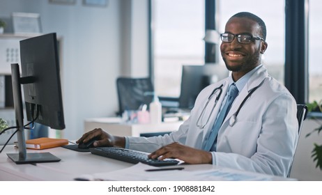 African American Family Medical Doctor In Glasses Is Working On A Computer In A Health Clinic. Successful Black Physician In White Lab Coat Looks At The Camera And Smiles In Hospital Office.